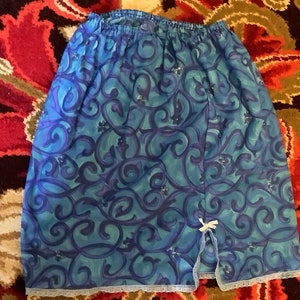May include: A blue and purple patterned slip with a white bow and lace trim. The slip has a side slit and an elastic waistband.