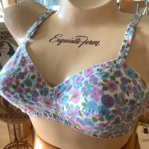 Puede incluir: Un sujetador blanco con estampado floral en tonos rosa, azul y morado. El sujetador tiene tirantes ajustables y cierre de gancho y ojo en la espalda. El sujetador está etiquetado como "Exquisite Form" en la parte delantera.