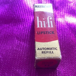 Könnte beinhalten: Ein gold- und roter Lippenstift-Behälter mit dem Text "hi-fi LIPSTICK AUTOMATIC REFILL" und "FROSTED ORANGE 32".