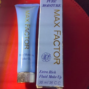 Könnte beinhalten: Eine blaue Tube Max Factor Pure Moisture Extra-Rich Fluid Make-Up mit silbernem Deckel. Die Tube liegt auf einer roten Oberfläche neben einer blauen Schachtel mit dem gleichen Produktnamen und dem Text "36 ml 36 C.C."