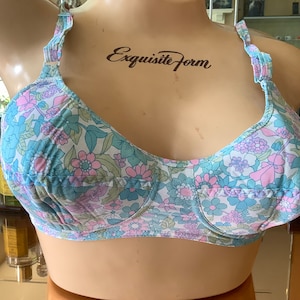 Puede incluir: Un sujetador de estampado floral con flores azules, rosas y verdes sobre un fondo blanco. El sujetador tiene tirantes ajustables y un cierre de gancho y ojo en la espalda. El sujetador está etiquetado como "Exquisite Form".