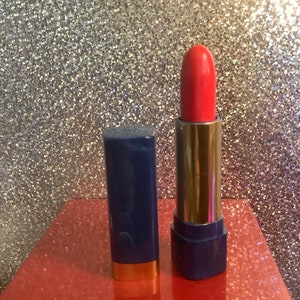 Könnte beinhalten: Ein roter Lippenstift in einem blauen und goldenen Röhrchen. Der Lippenstift ist geschlossen und das Röhrchen steht auf einer roten Oberfläche.