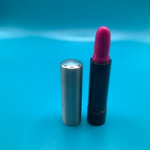 Könnte beinhalten: Ein pinkfarbener Lippenstift mit silberner Kappe und schwarzer Basis. Der Lippenstift hat eine leuchtende rosa Farbe und wird neben seiner silbernen Kappe gezeigt. Der Hintergrund ist einfarbig hellblau.