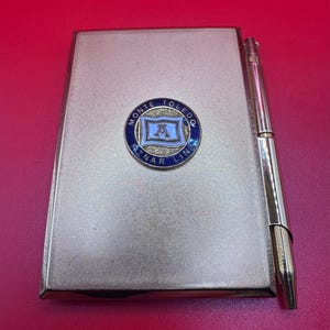 May include: A silver-toned cigarette case with a matching pen. The case has a textured surface and a circular emblem with the text "MONTE TOLEDO AZNAR LINE". The emblem is blue and white with a central design. The pen is positioned alongside the case.