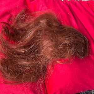 May include: A long, brown, wavy hair extension on a pink surface.