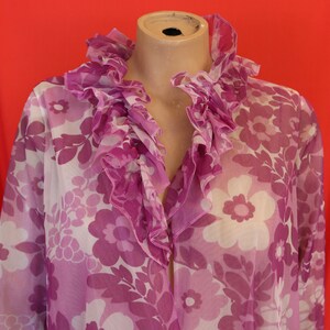 Könnte beinhalten: Eine durchsichtige, violette Bluse mit einem großen Rüschenkragen und einem Blumendruck. Die Blumen sind weiß mit violetten Zentren.