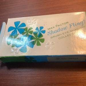 Può includere: Una scatola bianca con disegni di fiori stilizzati blu e verdi. La scatola è etichettata "Max Factor Shadow Fling! Brush Eye Shadow Collect."
