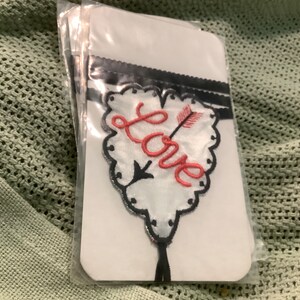 May include: White fabric patch with a heart-shaped design. The word "Love" is embroidered in coral-colored thread, with an arrow piercing the heart. The heart is outlined in black and has a scalloped edge. A black ribbon is at the top.