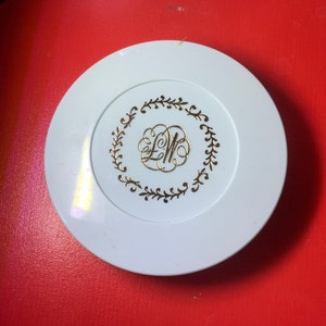 Könnte beinhalten: Ein runder weißer Kompakt mit einem goldenen Design, das ein Monogramm "LW" zeigt, das von einem Kranz aus Blättern umgeben ist.