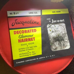 Könnte beinhalten: Eine rote runde Box mit einem Etikett, auf dem "Jacqueline Decorated Glamour Hairnet" in gelben Buchstaben steht. Das Etikett trägt außerdem die Aufschrift "styled by Jac-o-net" in schwarzen Buchstaben. Das Etikett hat ein kleines Fenster, das eine Probe von dunkelbraunem Haar zeigt.