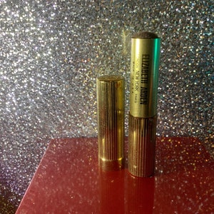 Könnte beinhalten: Zwei goldfarbene Kosmetikbehälter mit geripptem Design. Der Behälter rechts hat einen Puderapplikator und den Text "ELIZABETH ARDEN LONDON NEW YORK PARIS MADE IN U.S.A." auf der Seite.
