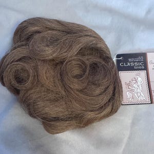 May include: A brown wig with tightly curled sections. The wig has a tag that reads "CLASSIC WIGS" with an illustration of two figures. The tag also states "100% HUMAN HAIR" and the color is "50 Tigette Gemini".
