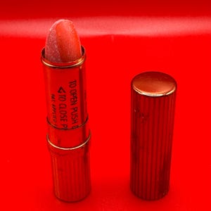 May include: A gold lipstick tube with a pink lipstick inside. The tube has the words "TO OPEN PUSH UP" and "TO CLOSE PUSH DOWN" printed on it.