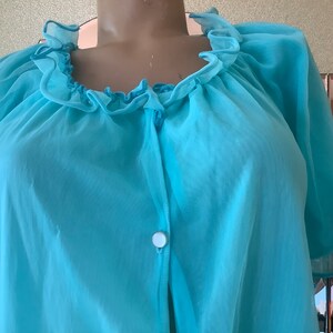 May include: A light blue vintage chiffon robe with a ruffled neckline and sleeves. The robe has a button closure and is made of a sheer, lightweight fabric. The sleeves are short and flared.