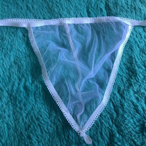 Könnte beinhalten: Weißer, durchsichtiger String-Tanga mit zarten, gewellten Verzierungen. Die Dessous haben ein dreieckiges Design und einen dünnen Bund. Der Stoff ist leicht und durchscheinend und erzeugt eine minimalistische Ästhetik.