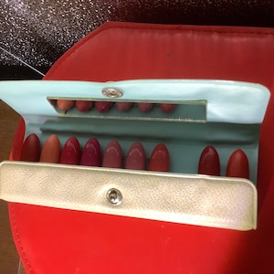 Op de afbeelding: Een vintage lippenstift etui met een lichtblauwe buitenkant en een witte binnenkant. Het etui heeft een spiegel en bevat tien lippenstift buisjes.