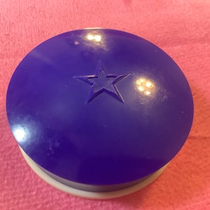 May include: A round, purple compact with a white star design on the lid. The compact is made of plastic and has a white base.