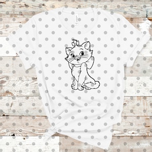 May include: White t-shirt featuring a black and white illustration of a cartoon cat with a bow and a bow on its head. The shirt has a crew neck and short sleeves. The background is a light gray polka dot pattern.