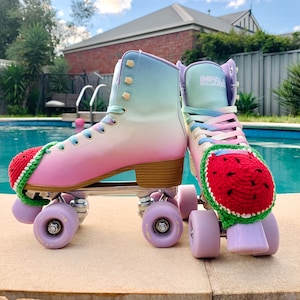 May include: Pair of Impala roller skates with a gradient color scheme of pink, white, and blue. The skates have purple wheels and are adorned with red, green, and white crocheted watermelon toe caps. The laces are pastel rainbow.