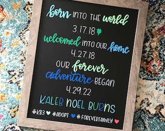 May include: A black chalkboard with white text announcing the birth of a child. The text reads "born into the world 3.17.18", "welcomed into our home 4.27.18", "our forever adventure began 4.29.22", "Kaleb Noel Burns", "#KB3 #ADOPT #FOREVERFAMILY".