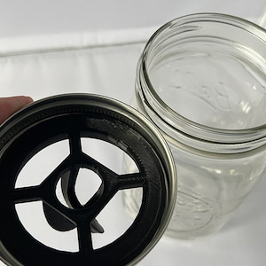 May include: A clear glass mason jar with a silver metal ring lid featuring a black, circular, 3D-printed insert with a cross-shaped support structure and a central opening, designed to fit securely within the mouth of the jar.