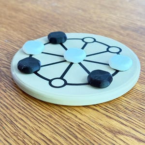 May include: A beige board game with a circular design and black lines. The game includes black and light blue game pieces. The board is on a wooden surface.