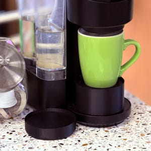 May include: A black coffee maker with a green mug on the drip tray. The coffee maker has a clear water reservoir and a black lid. There is a jar of coffee pods in the foreground.