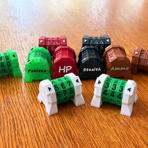 May include: A selection of colourful, 3D-printed game dice in green, red, black, brown, and white. Some dice display numbers, while others are labelled with terms such as "Potions," "HP," "Stealth," and "Ammo." The dice are arranged on a wooden surface.
