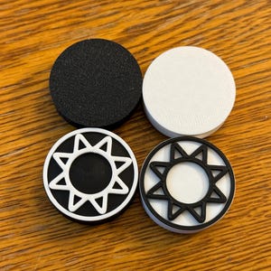 May include: Four round objects are arranged on a wooden surface. Two are solid: one black and textured, one white and smooth. The other two feature a sunburst design, one with a white sun on a black background, the other with a black sun on a white background.