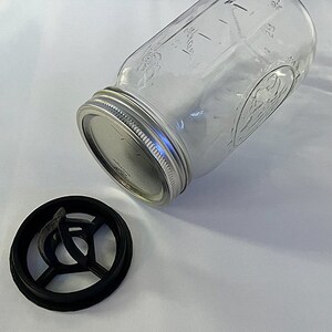 May include: A clear glass mason jar with a silver metal lid and a separate black plastic circular insert featuring a cross-shaped support structure. The jar has embossed text and markings on its surface, and the lid is securely screwed on.