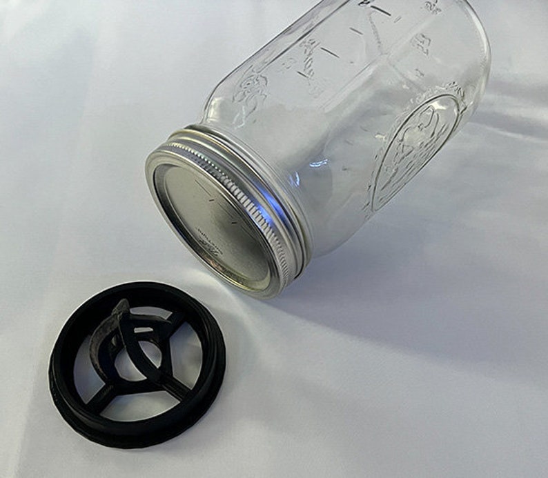 May include: A clear glass mason jar with a silver metal lid and a separate black plastic circular insert featuring a cross-shaped support structure. The jar has embossed text and markings on its surface, and the lid is securely screwed on.