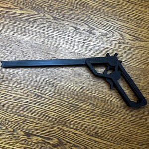 May include: A black plastic toy gun with a long barrel and a handle. The gun is lying on a wooden surface.