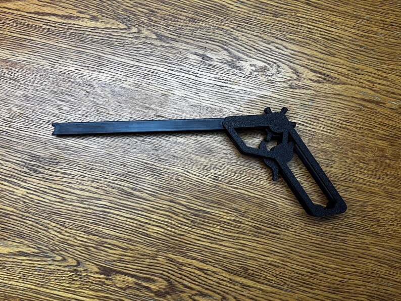 May include: A black plastic toy gun with a long barrel and a handle. The gun is lying on a wooden surface.