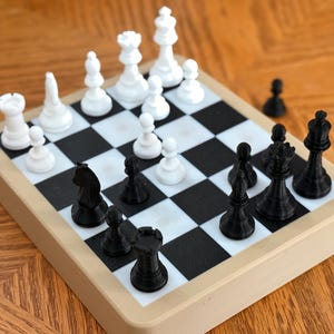 May include: A miniature chess set with black and white pieces arranged on a checkered board. The board is set on a light tan frame, and the pieces are in the starting positions. The chess set is on a wooden surface.
