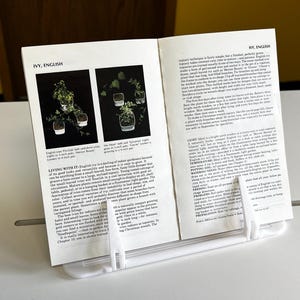 May include: Two pages from a book about gardening, showing close-up photos of Ivy English plants in pots. The text on the page describes the plant's care and characteristics. The book is open and propped up on a white book stand.