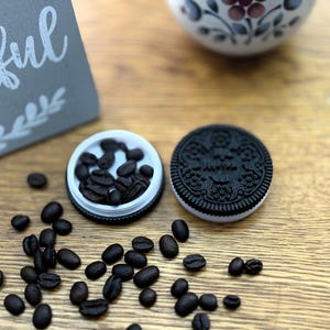 May include: A white container with a black lid filled with coffee beans. A black and white cookie with a decorative design is next to the container. Coffee beans are scattered on a wooden surface.