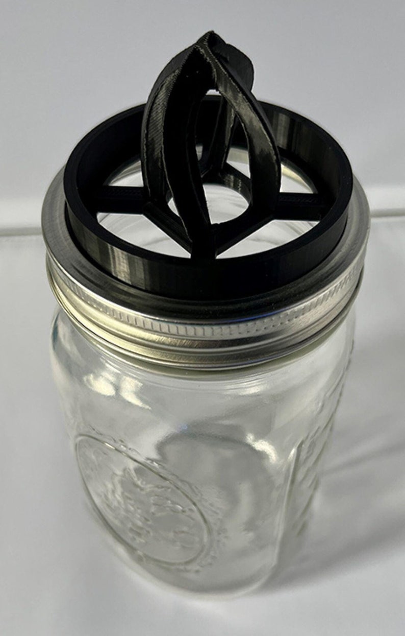 May include: A clear glass mason jar featuring a silver metal screw-top ring and a black plastic insert. The insert has a circular frame with a central, arched, multi-ribbed structure extending upward from the opening of the jar.