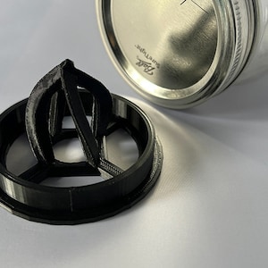May include: A black 3D-printed citrus juicer attachment designed to fit onto a glass jar. The circular base features a central reamer with three vertical fins. Beside it sits a silver metal Ball SureTight lid for a standard mason jar.