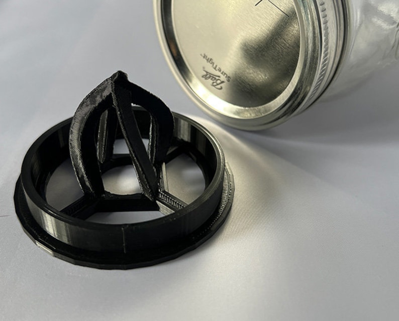 May include: A black 3D-printed citrus juicer attachment designed to fit onto a glass jar. The circular base features a central reamer with three vertical fins. Beside it sits a silver metal Ball SureTight lid for a standard mason jar.