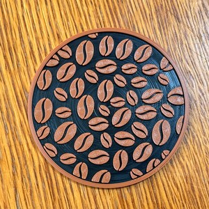 May include: A round, coffee-themed coaster with a black center and a brown border. The center features a pattern of brown coffee beans. The coaster is designed to protect surfaces from hot or cold beverages.