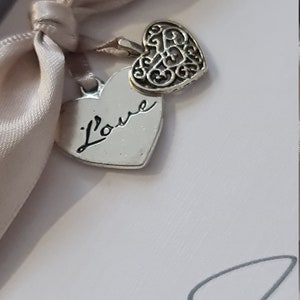 May include: A white box with a beige satin ribbon tied around it. Two silver heart charms hang from the ribbon. One charm has the word "l'ove" inscribed on it, and the other charm has a decorative filigree design.