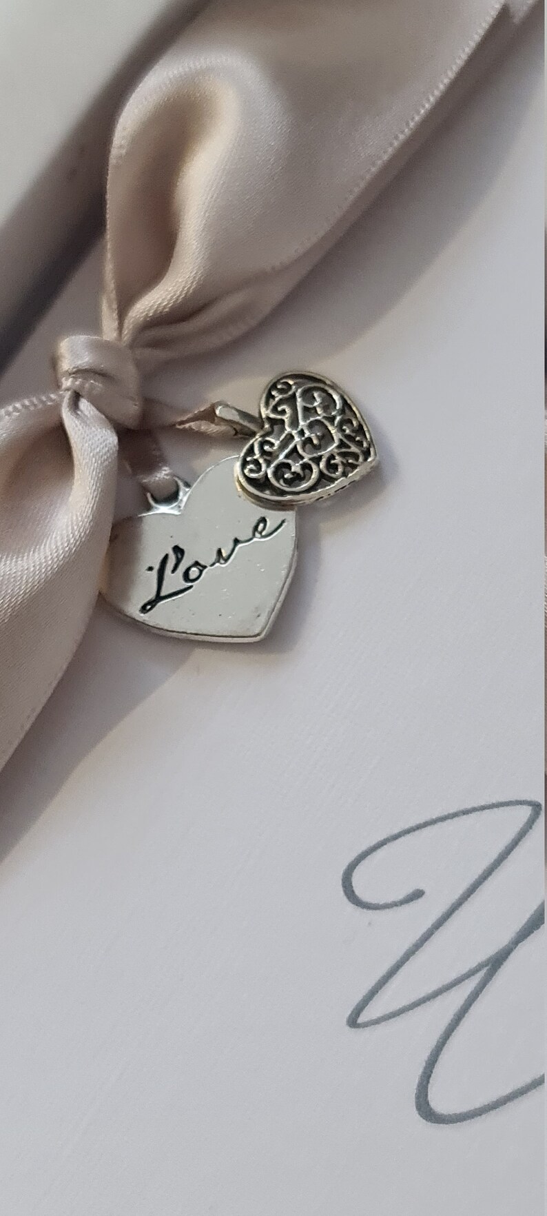 May include: A white box with a beige satin ribbon tied around it. Two silver heart charms hang from the ribbon. One charm has the word "l'ove" inscribed on it, and the other charm has a decorative filigree design.