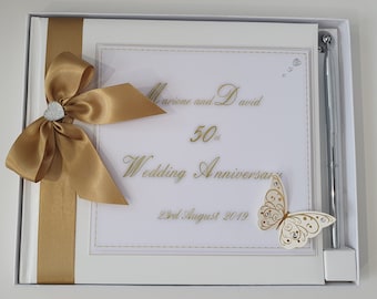 May include: White wedding guest book with a gold ribbon and bow, a gold pen, and a gold butterfly with rhinestones. The book has the text "Mariene and David 50th Wedding Anniversary 22nd August 2019" on the front.