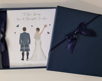Puede incluir: Una caja de regalo azul marino con una tarjeta blanca en su interior. La tarjeta presenta una ilustración de dibujos animados de una novia y un novio. El texto de la tarjeta dice "To Our Lovely Son & Daughter In Law, Christopher and Chloe".