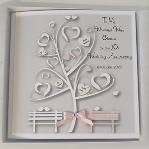Puede incluir: Una tarjeta blanca con un fondo gris que presenta un árbol de papel recortado blanco con corazones rosas y un banco con una cinta rosa. La tarjeta dice "To My Wonderful Wife Charlene On Our 10th Wedding Anniversary 8th October 2020".