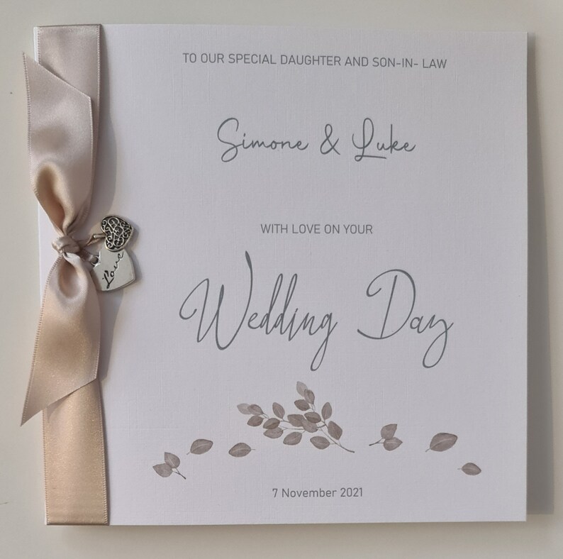 May include: A white card with a beige ribbon and a silver charm that says "Love". The card has the text "To our special daughter and son-in-law Simone & Luke With love on your Wedding Day 7 November 2021".