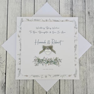 May include: A white card with a green floral design and a silver heart charm with the words "Mr & Mrs" on it. The card says "Wedding Day Wishes To Our Daughter & Son In Law Hannah & Robert 24.06.2023".