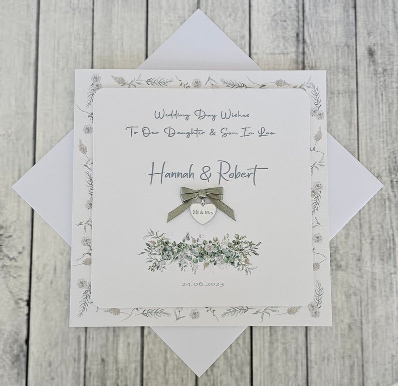 May include: A white card with a green floral design and a silver heart charm with the words "Mr & Mrs" on it. The card says "Wedding Day Wishes To Our Daughter & Son In Law Hannah & Robert 24.06.2023".
