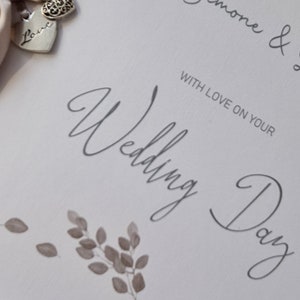 May include: A white card with the text "To our special daughter Simone & with love on your Wedding Day 7 November 2021". The card is decorated with a beige satin ribbon and two silver charms, one with the word "Love" and the other with a decorative heart design.