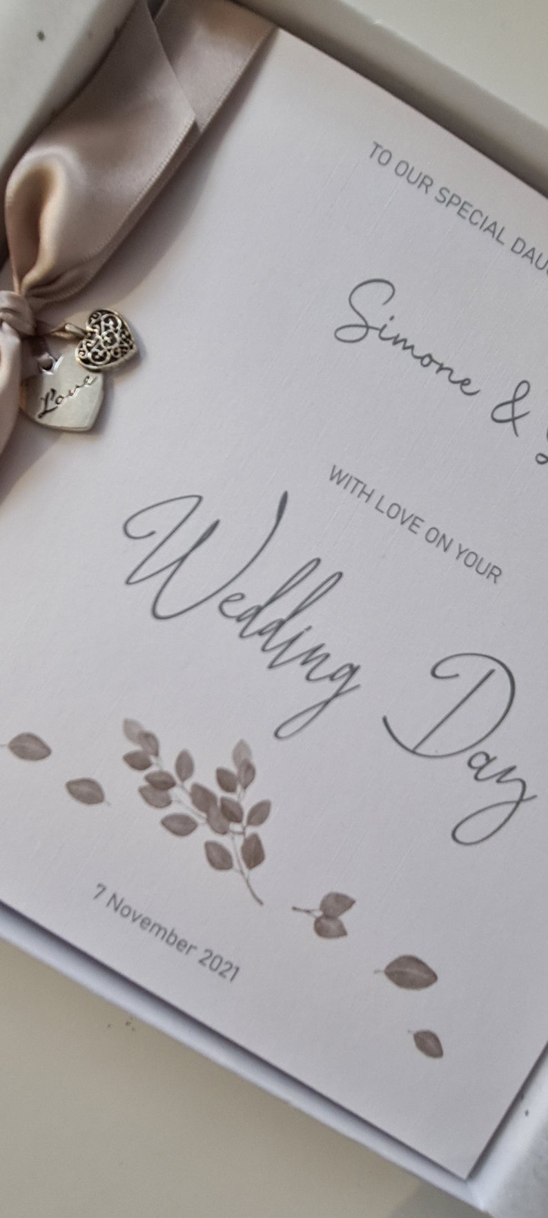May include: A white card with the text "To our special daughter Simone & with love on your Wedding Day 7 November 2021". The card is decorated with a beige satin ribbon and two silver charms, one with the word "Love" and the other with a decorative heart design.